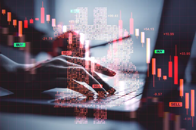 Ein roter Bitcoin-Kurs, dahinter arbeitet jemand an einem Laptop.