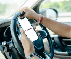 Auf dem Bild ist der Innenraum eines Autos zu sehen. Eine Person sitzt am Steuer und hält mit einer Hand das Lenkrad, während sie mit der anderen ein Smartphone in der Hand hat. Auf dem Display des Smartphones ist das TomTom-Logo zu sehen.
