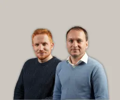 Auf dem Bild sind zwei Männer vor neutralem Hintergrund zu sehen: Links steht Georg Buschmann und rechts Henry Philippson. Beide blicken direkt in die Kamera.